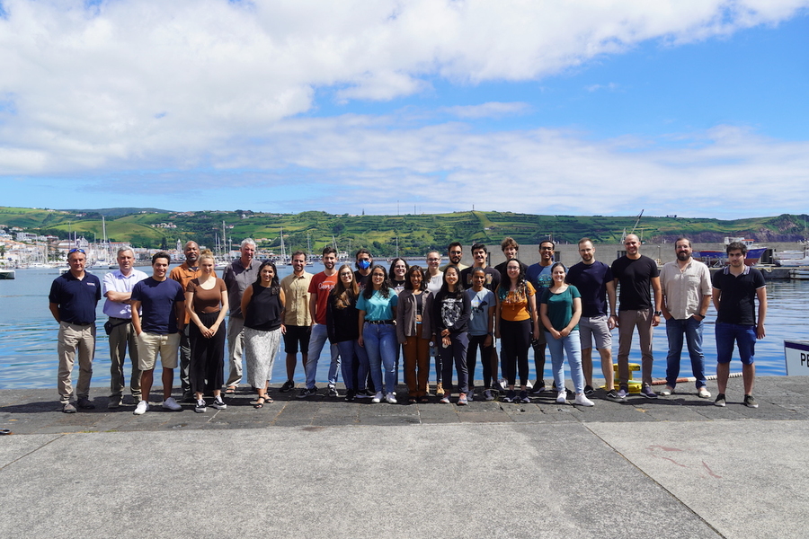 A group of people standing side by side pose for a photo on a concrete pier, with water behind them. Low hills are in the background.