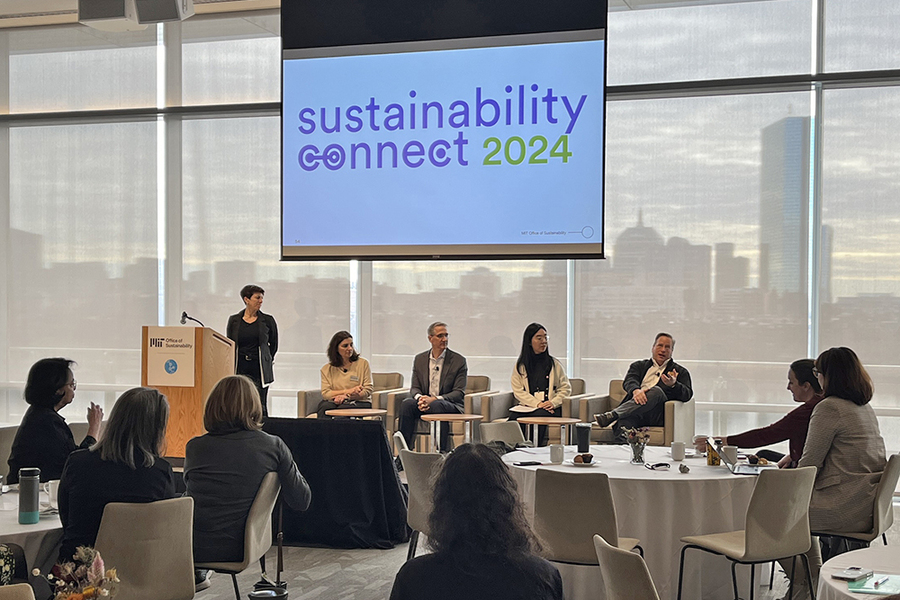 One person stands behind a lectern with four seated panelists to her left. Above them, a screen displays "Sustainability connect 2024." Boston’s skyline fills the windows behind them.