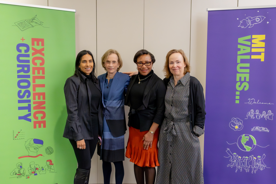 Four women pose in a line against a wall, flanked by banners reading “Excellence Curiosity” and “MIT Values."