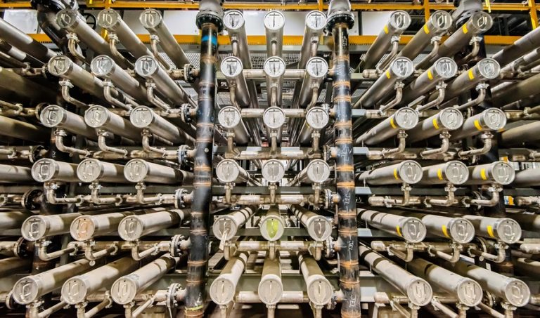 A rack of interconnected large silver tubes and pipes used to house filtration membranes for industrial chemical processing.