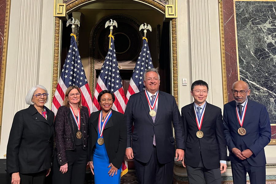 Arati Prabhakar, Angela Belcher, Paula Hammond, Noubar Afeyan, Feng Zhang, and Emory Brown pose together in front of 3 U.S. flags.